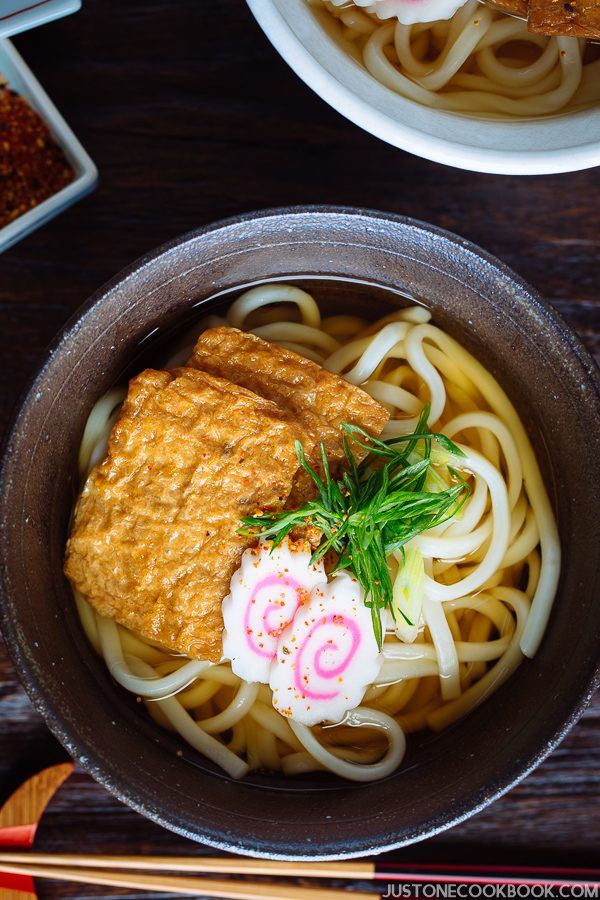 A dark bowl containing udon noodles in dashi broth topped with deep fried tofu, fish cake, green onion, and sprinkle of shichimi togarashi on the table.