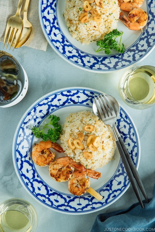 A blue and white plate containing Japanese Garlic Fried Rice topped with garlic chips and grilled shrimp skewer.