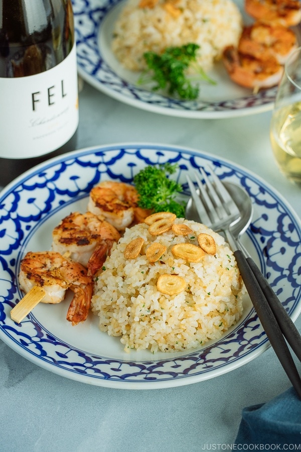 A blue and white plate containing Japanese Garlic Fried Rice topped with garlic chips and grilled shrimp skewer. Served with a glass of wine.