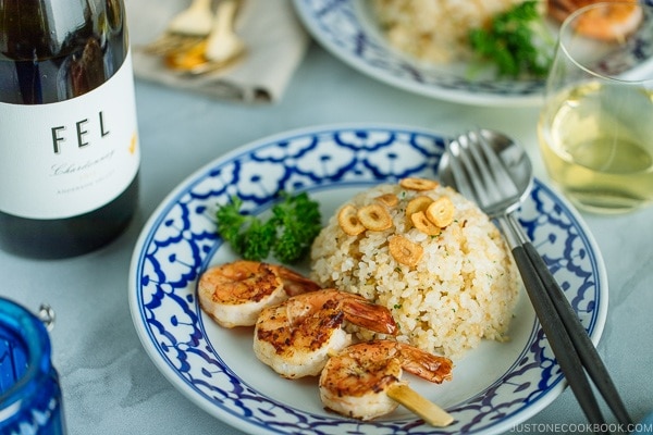 A blue and white plate containing Japanese Garlic Fried Rice topped with garlic chips and grilled shrimp skewer. Served with a glass of wine.