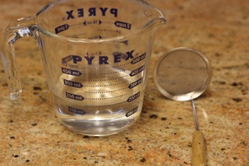 water in a measuring cup next to a mesh skimmer on granite countertop