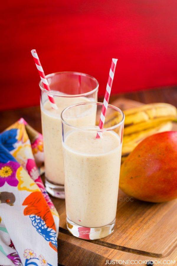 Two glasses of Mango Coconut Smoothie, banana and mango on a wooden cutting board.