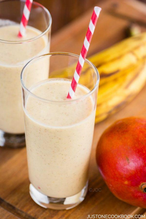 Two glasses of Mango Coconut Smoothie, banana and mango on a table.