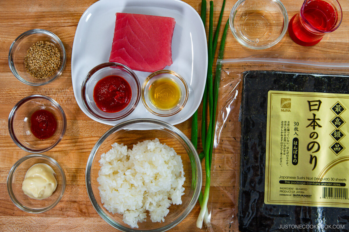 A top-down view of sushi ingredients on a wooden surface: a block of raw tuna, sushi rice, nori sheets, green onions, sesame seeds, sauces in small bowls, and mayonnaise&mdash;perfect for making a spicy tuna roll.