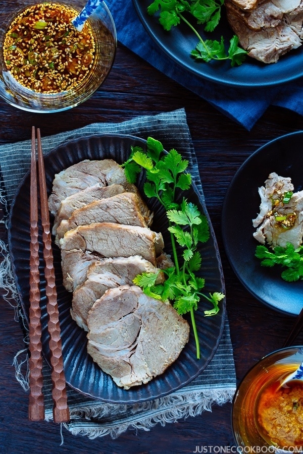 A dark plate containing sliced Pressure Cooker Steamed Pork, surrounded by 2 glass bowls, each of which holds garlic soy sauce and ginger miso sauce.