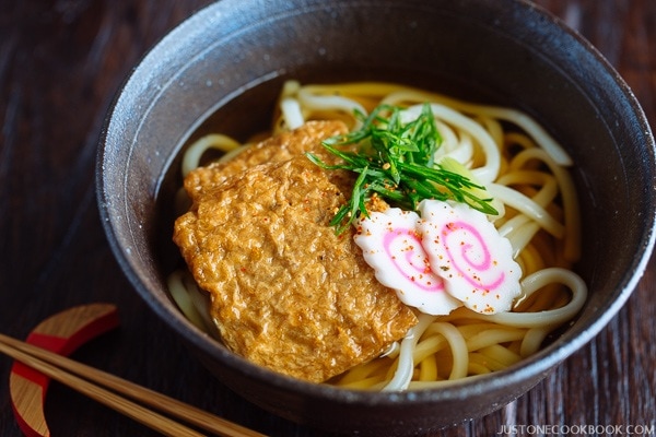 A dark bowl containing udon noodles in dashi broth topped with deep fried tofu, fish cake, green onion, and sprinkle of shichimi togarashi.