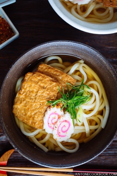 A dark bowl containing udon noodles in dashi broth topped with deep fried tofu, fish cake, green onion, and sprinkle of shichimi togarashi.