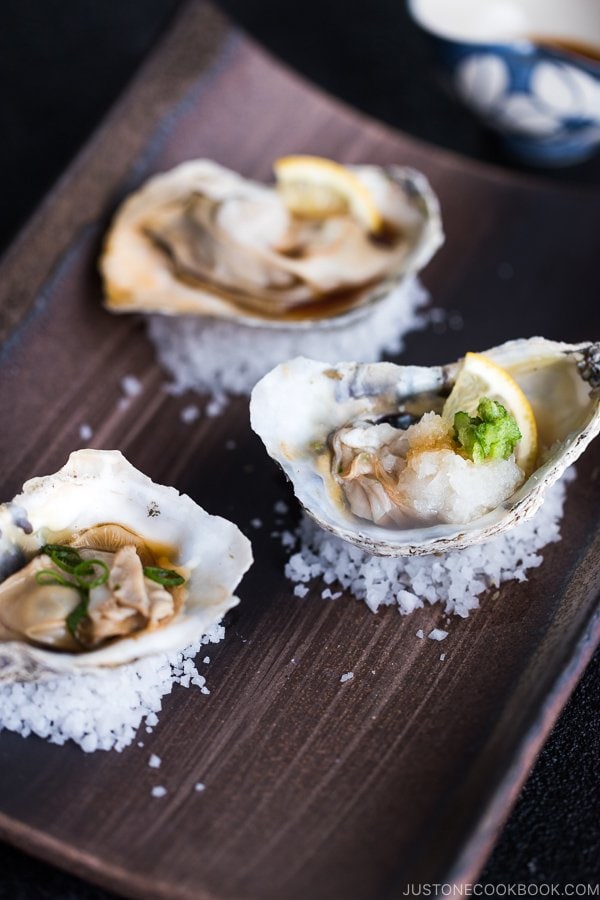 Dark Japanese plate containing Grilled Oysters with 3 kinds of garnish and Ponzu Sauce