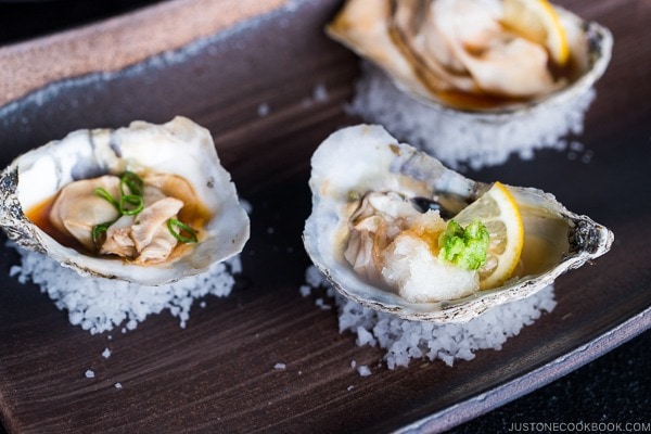 Dark Japanese plate containing Grilled Oysters with 3 kinds of garnish and Ponzu Sauce