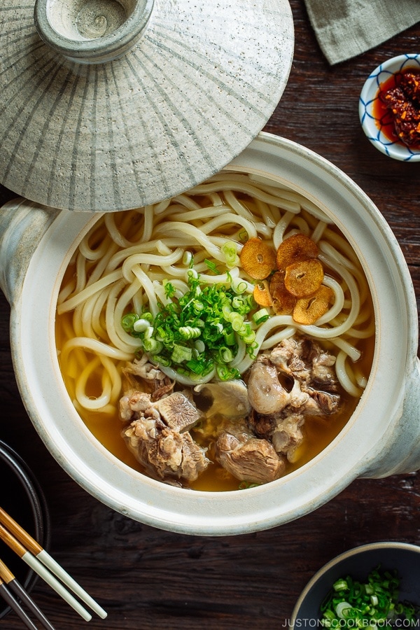 A Japanese donabe (earthenware pot) containing Oxtail Udon topped with tender meat, fried garlic, and green onion.