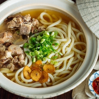 A Japanese donabe (earthenware pot) containing Oxtail Udon topped with tender meat, fried garlic, and green onion.