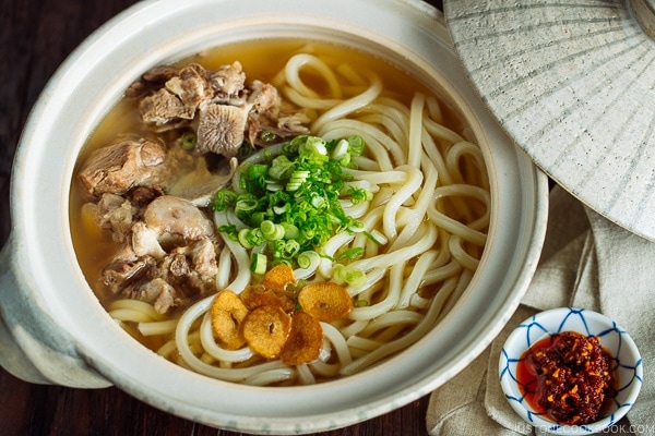 A Japanese donabe (earthenware pot) containing Oxtail Udon topped with tender meat, fried garlic, and green onion.