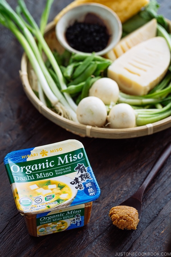 A tub of "Organic Miso - Dashi Miso" by Hikari Miso. A bamboo basket behind containing vegetables.