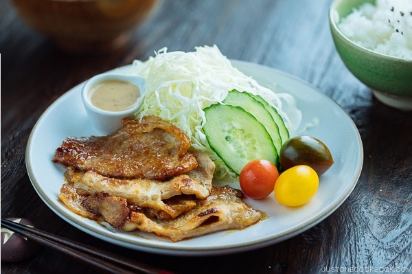 A white plate containing thinly sliced Miso Ginger Pork served with shredded cabbage, sliced cucumbers, and cherry tomatoes.