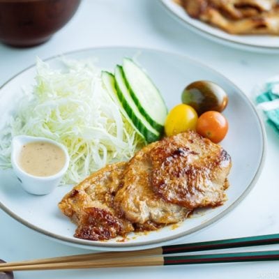 A white plate containing thinly sliced Miso Ginger Pork served with shredded cabbage, sliced cucumbers, and cherry tomatoes.