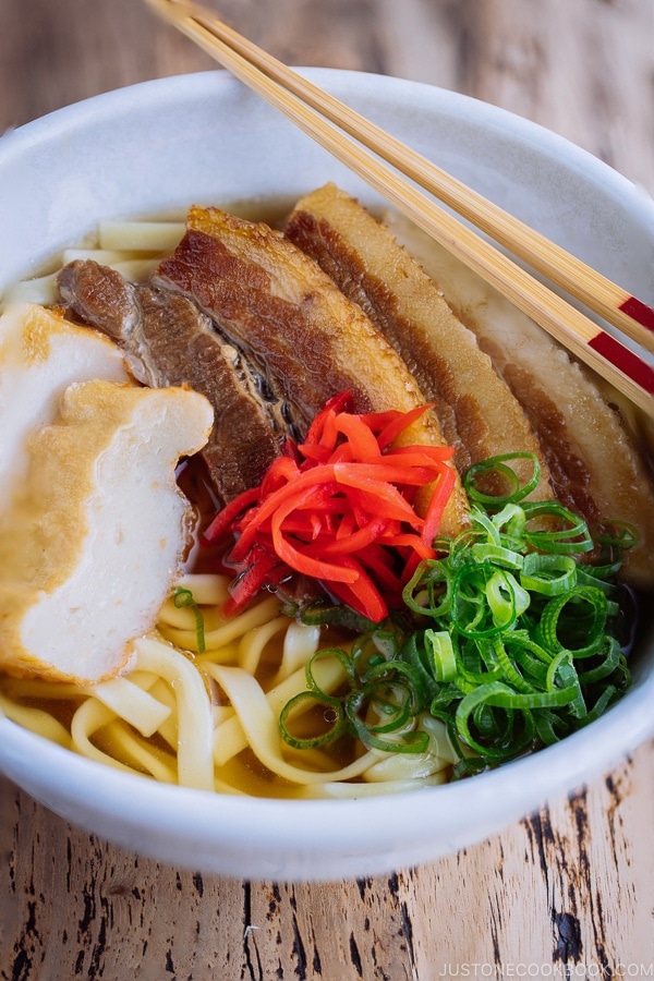 A white bowl containing Okinawa Soba and delicious Japanese dashi and pork broth, topped braised pork belly, fish cakes, green onion, and red pickled ginger.