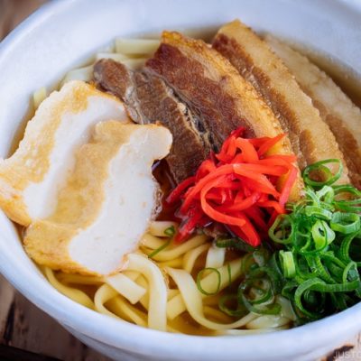 A white bowl containing Okinawa Soba and delicious Japanese dashi and pork broth, topped braised pork belly, fish cakes, green onion, and red pickled ginger.