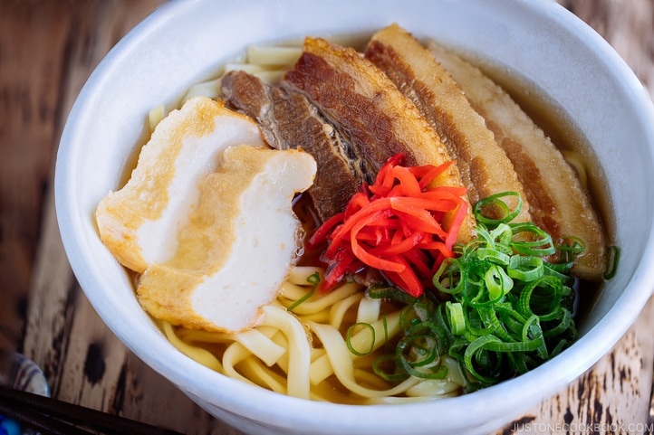 A white bowl containing Okinawa Soba and delicious Japanese dashi and pork broth, topped braised pork belly, fish cakes, green onion, and red pickled ginger.