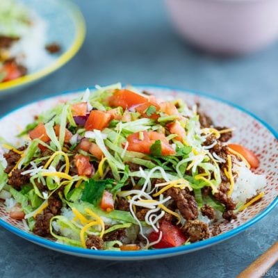 Okinawa Taco Rice on a colorful plate consisting of white rice, taco seasoning ground beef, shredded lettuce and cheese, topped with tomato salsa.