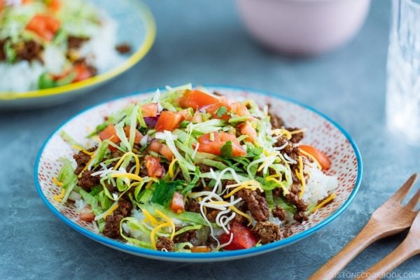 Okinawa Taco Rice on a colorful plate consisting of white rice, taco seasoning ground beef, shredded lettuce and cheese, topped with tomato salsa.
