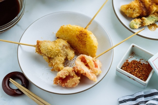 A white plate containing golden and crispy Kushikatsu (Kushiage) served with shichimi togarashi (Japanese seven spice).