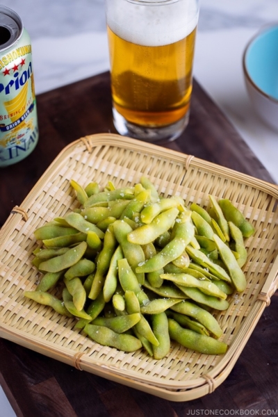 Edamame served in bamboo basket and beer glass in the back.