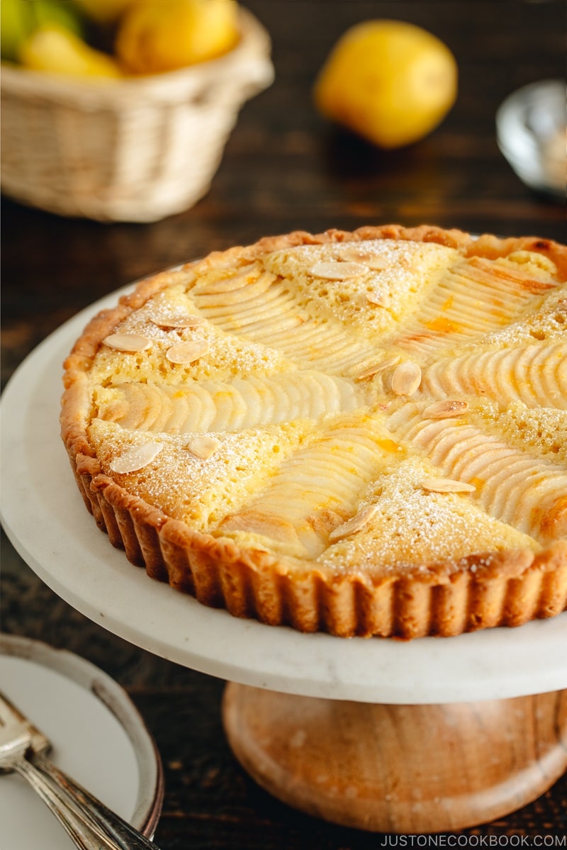 Pear and Almond Tart (Pear Frangipane Tart) is on the cake stand. The tart is dusted with powdered sugar and sprinkled with toasted almond slices.