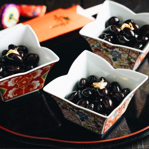 Sweet black soybeans garnished with a gold leaf in Japanese red and gold dishes.
