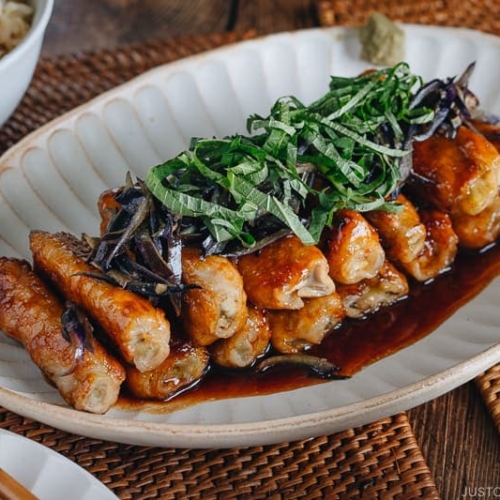 A Japanese white plate containing ginger pork rolls with eggplant garnished with shiso leaves on top.