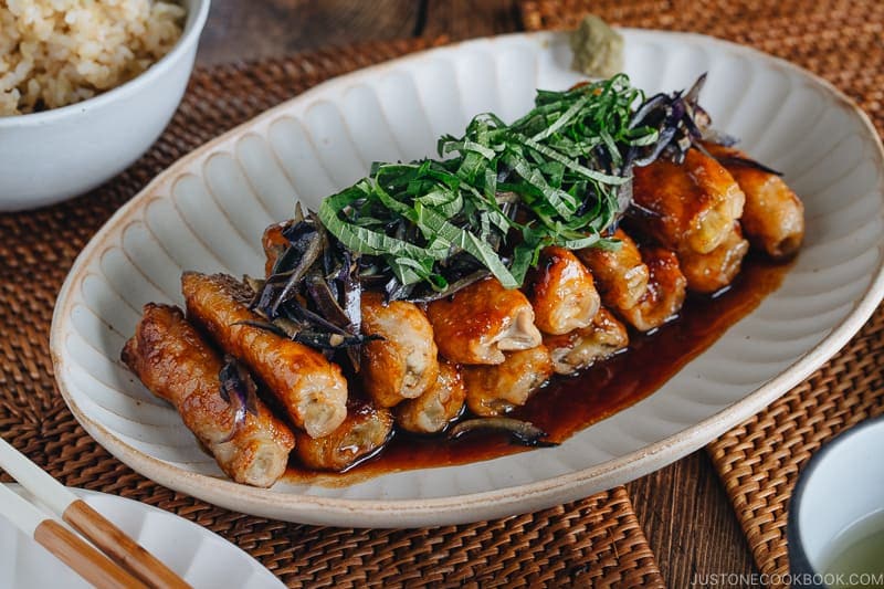 A Japanese white plate containing ginger pork rolls with eggplant garnished with shiso leaves on top.