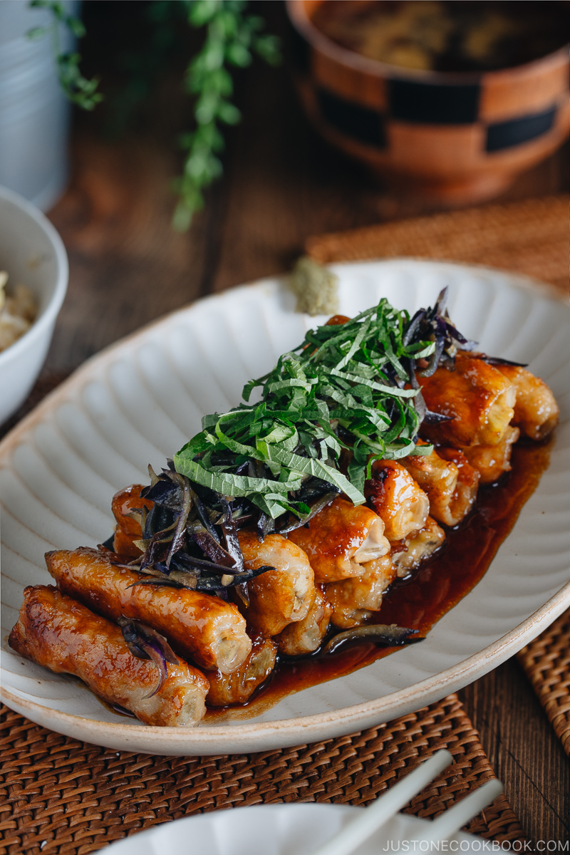 A Japanese white plate containing ginger pork rolls with eggplant garnished with shiso leaves on top.