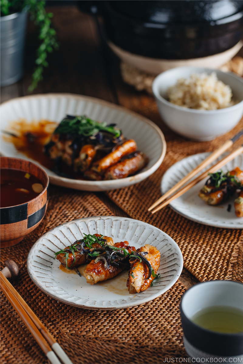 A Japanese white plate containing ginger pork rolls with eggplant garnished with shiso leaves on top.