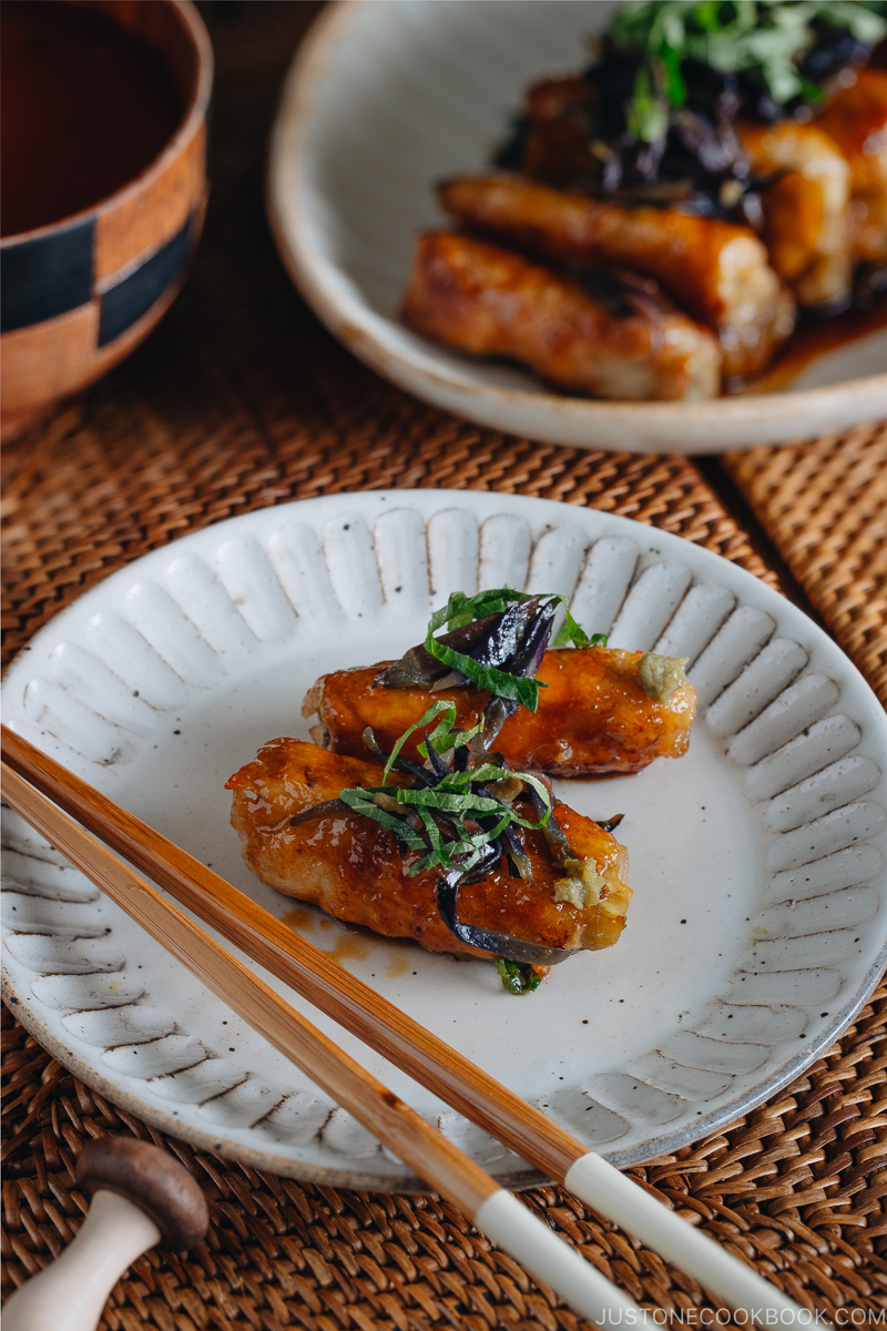 A Japanese white plate containing ginger pork rolls with eggplant garnished with shiso leaves on top.