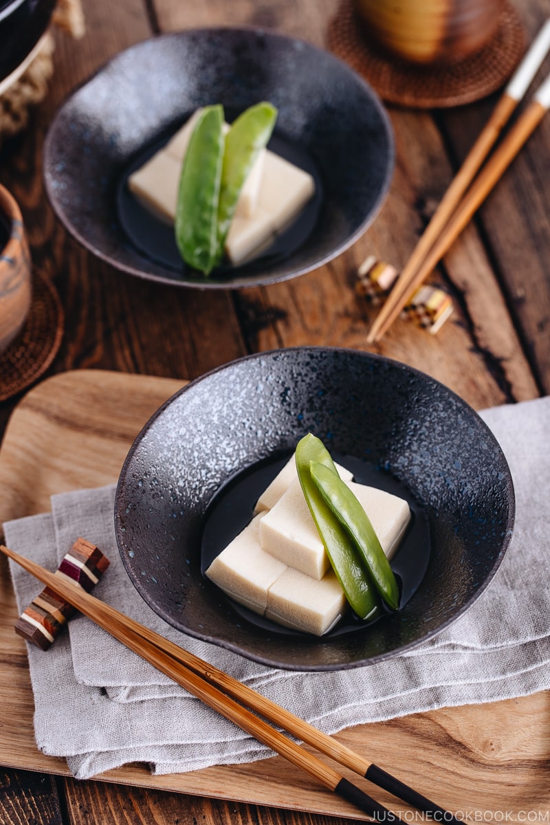 Simmered koyadofu with snow peas in a black ceramic bowl.