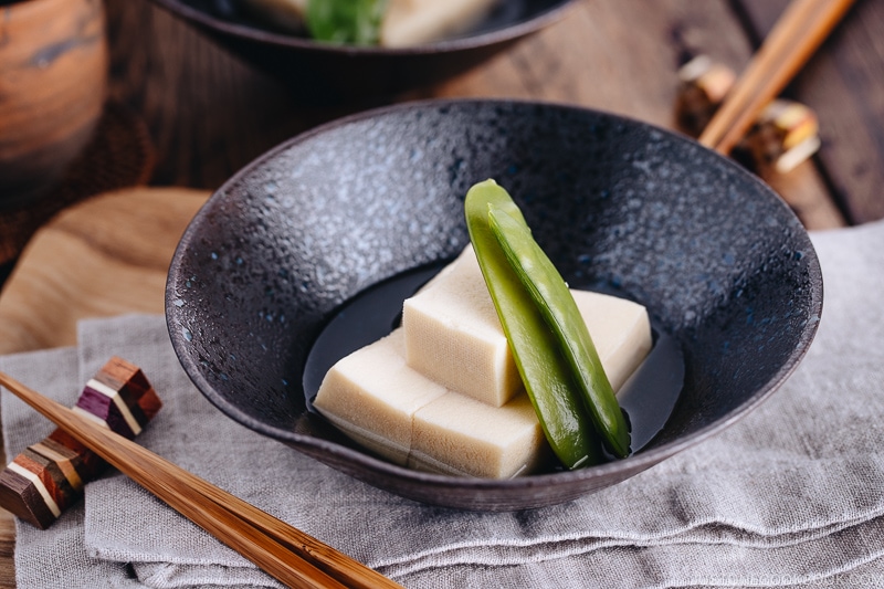 Simmered koyadofu with snow peas in a black ceramic bowl.