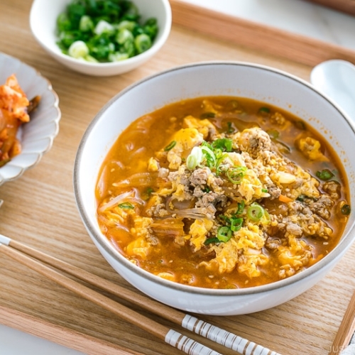 A white bowl containing kimchi soup that's made of Malony glass noodles, kimchi, egg, and pork.