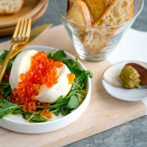 A white plate containing arugula and burrata cheese topped with ikura and served with baguette and yuzu kosho pepper.