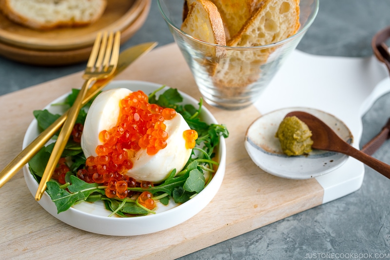 A white plate containing arugula and burrata cheese topped with ikura and served with baguette and yuzu kosho pepper.