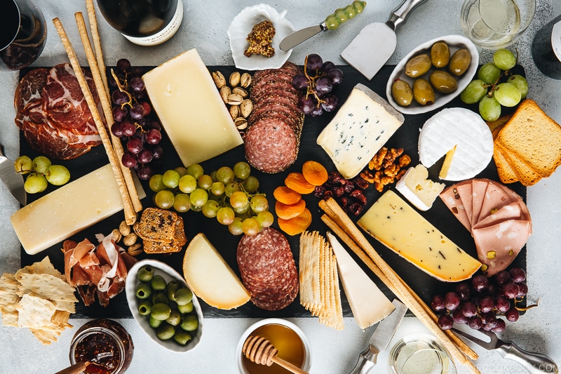 Cheese board filled with cheese, cold cuts, dried fruits, bread sticks, olives, grapes, and condiments.