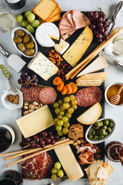 Cheese board filled with cheese, cold cuts, dried fruits, bread sticks, olives, grapes, and condiments.