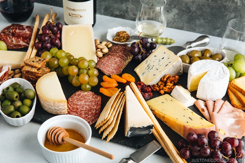 Cheese board filled with cheese, cold cuts, dried fruits, bread sticks, olives, grapes, and condiments.