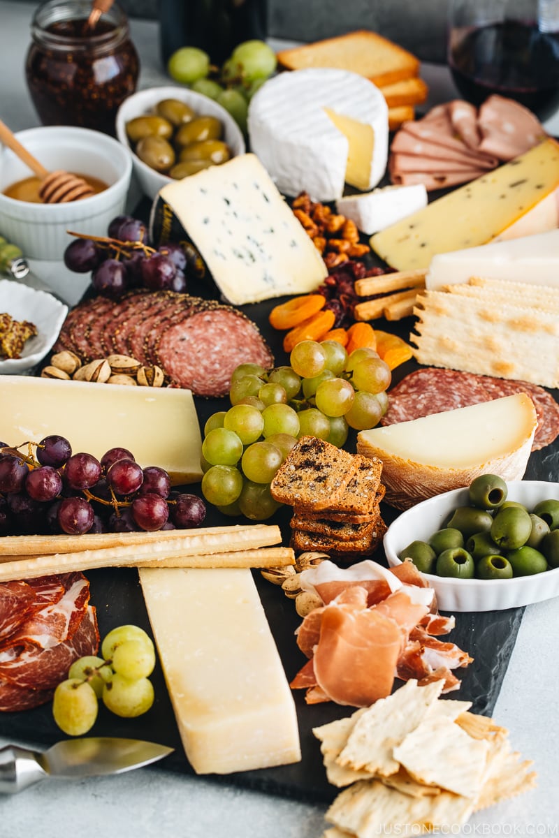 Cheese board filled with cheese, cold cuts, dried fruits, bread sticks, olives, grapes, and condiments.