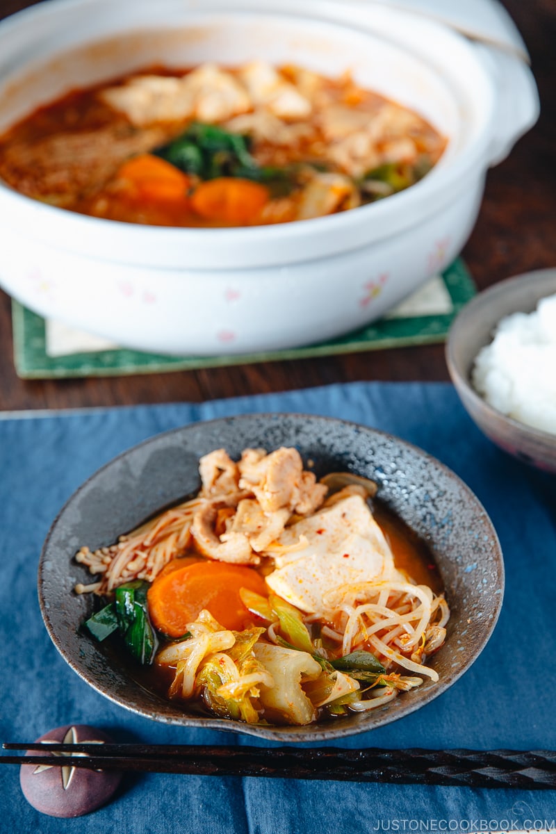 A black ceramic bowl containing kimchi stew along with a big pot of kimchi stew.