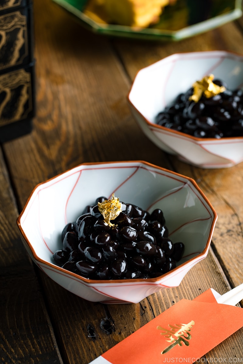 A white and red Japanese bowl containing Kuromame, sweet black soybeans, topped with gold leaf for the Japanese new year's celebration.