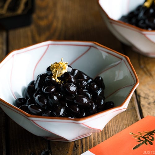 A white and red Japanese bowl containing Kuromame, sweet black soybeans, topped with gold leaf for the Japanese new year's celebration.