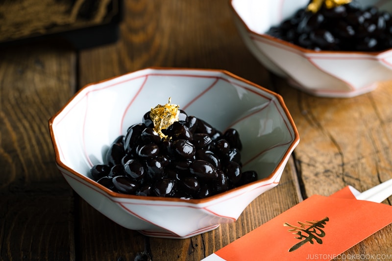 A white and red Japanese bowl containing Kuromame, sweet black soybeans, topped with gold leaf for the Japanese new year's celebration.