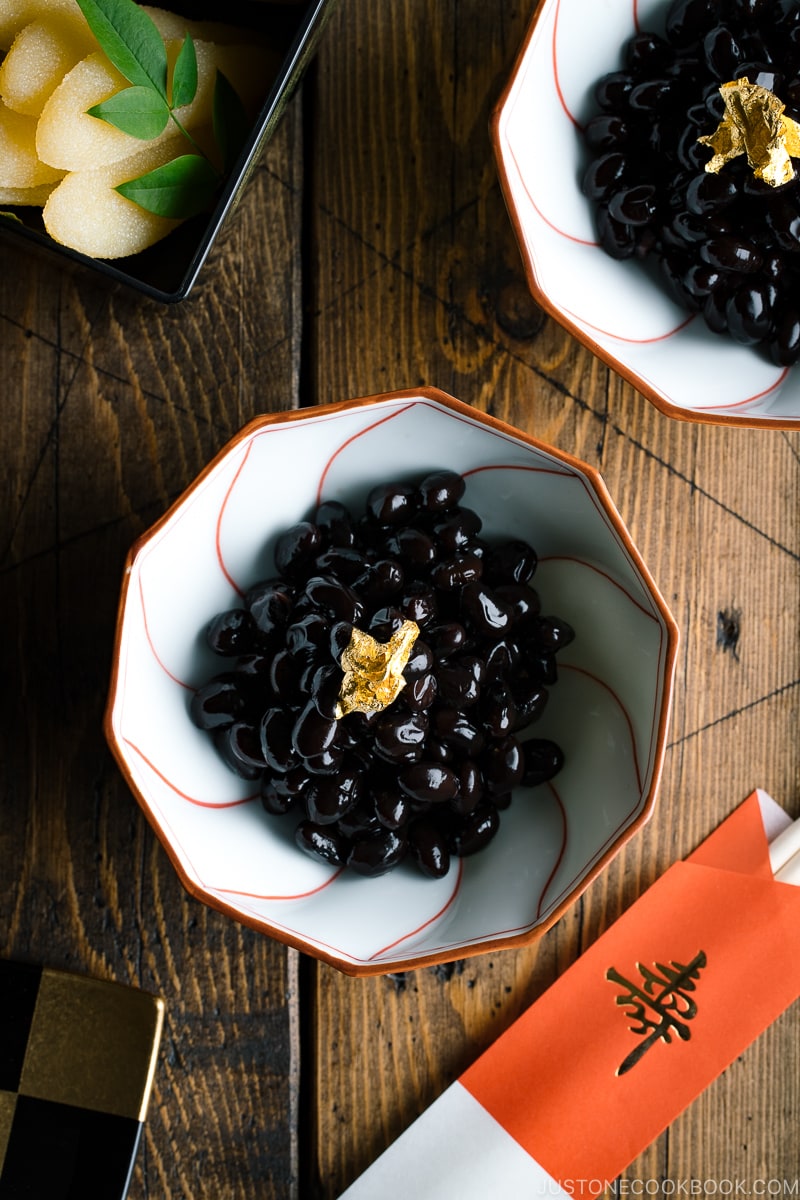 A white and red Japanese bowl containing Kuromame, sweet black soybeans, topped with gold leaf for the Japanese new year's celebration.