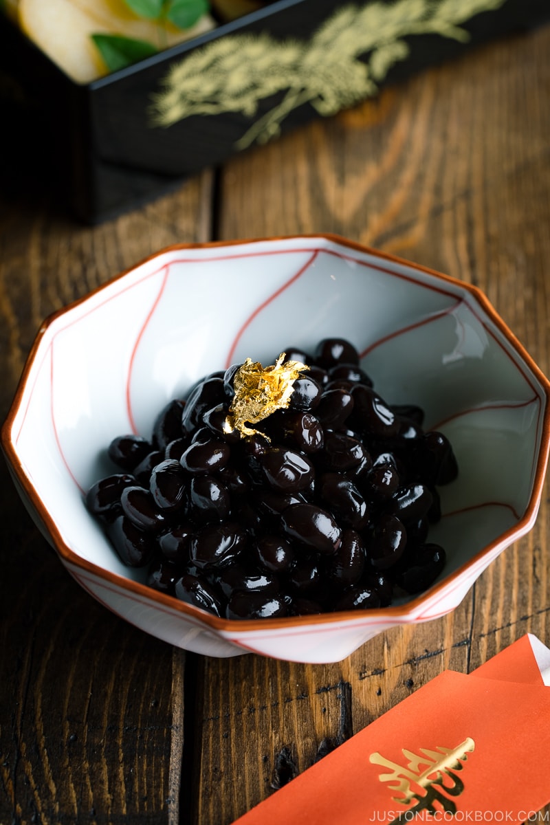 A white and red Japanese bowl containing Kuromame, sweet black soybeans, topped with gold leaf for the Japanese new year's celebration.