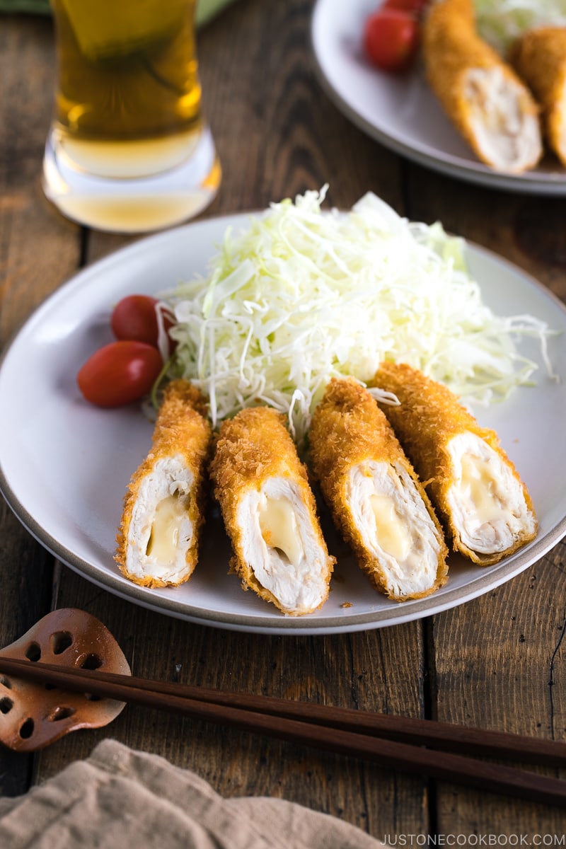 A white ceramic plate containing chicken cheese katsu served with shredded cabbage and cherry tomato.