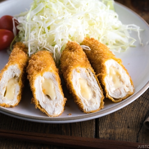 A white ceramic plate containing chicken cheese katsu served with shredded cabbage and cherry tomato.